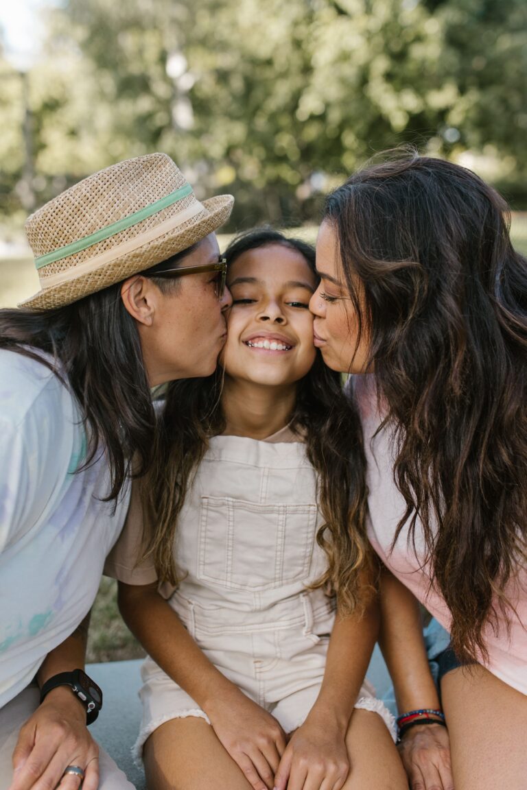 Two mothers kissing their young daughter on the cheeks outdoors